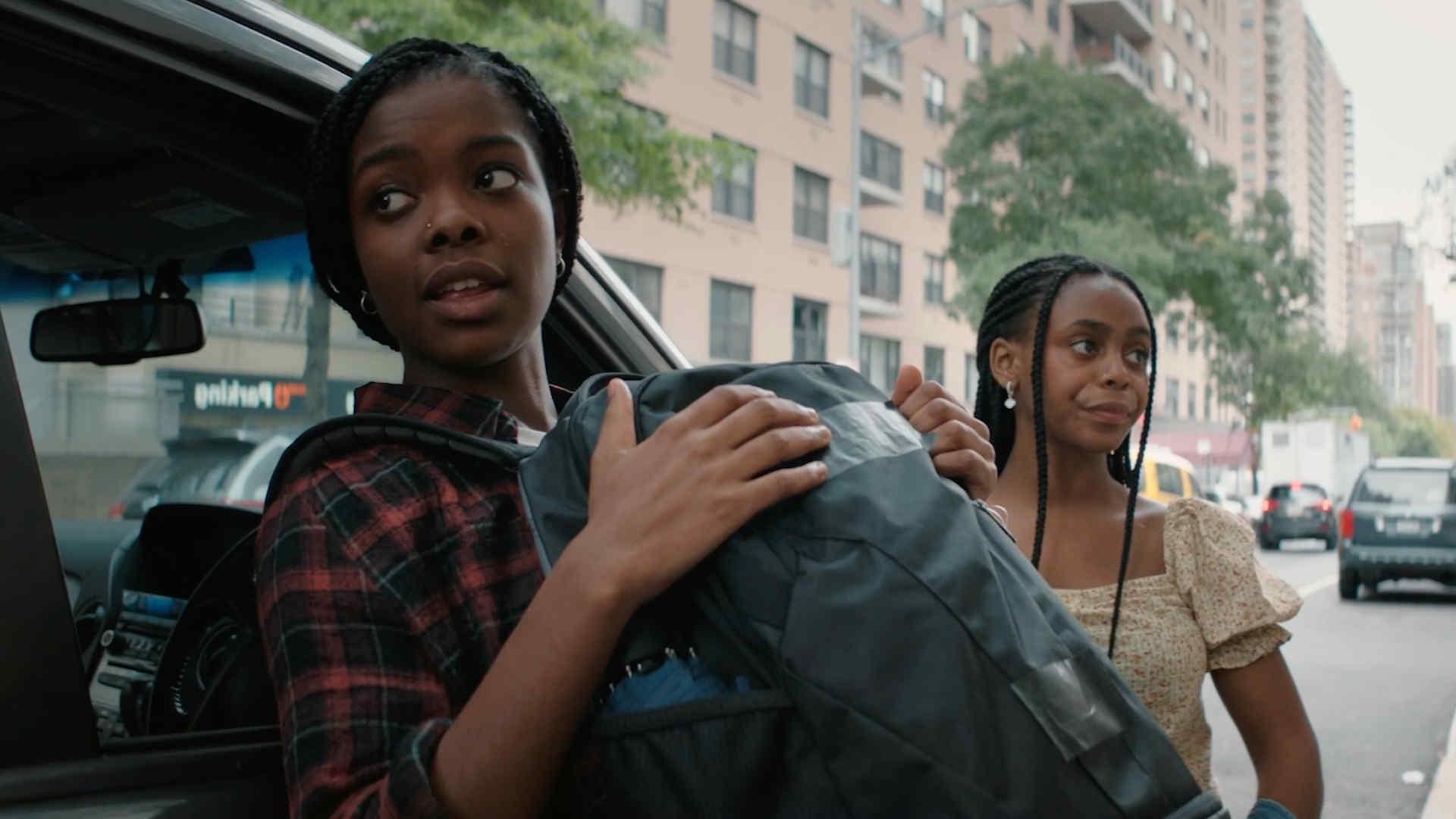 In this still from the film, two girls are exiting a car onto a busy Manhattan street. One girl is holding a backpack and they have facial expressions of concern